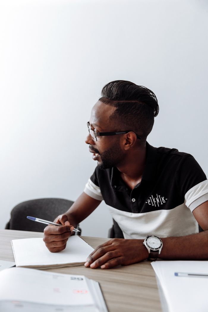Man sitting at a desk writing in an office environment, showcasing concentration and style.