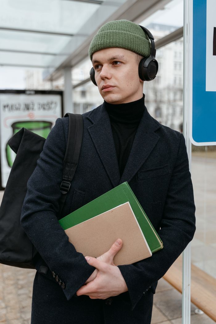 about-img Man in beanie with headphones, holding books at an outdoor transit station.