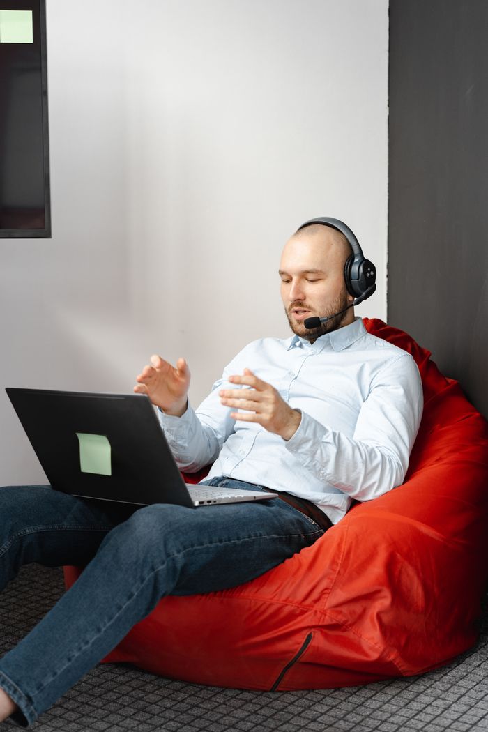 gallery-img-04 Man working remotely on laptop with headset, seated on a red beanbag chair in a modern indoor setting.